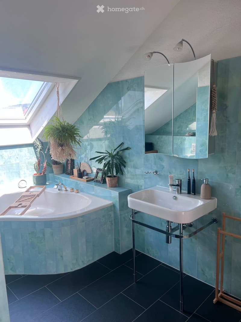 Modern bathroom with blue tiles, a bathtub, a sink, a large mirror, and decorative plants on the windowsill.
