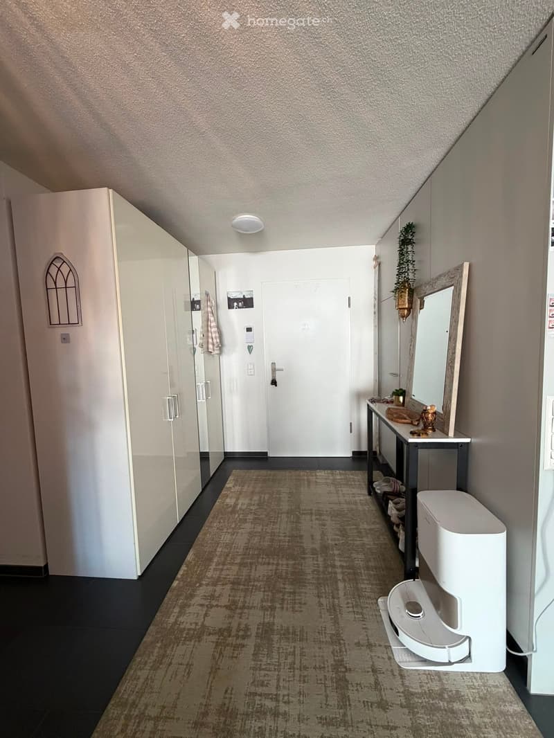 Hallway with carpeted floor, white walls, a small table with a mirror, and a white trash bin. There is a door at the end of the hallway.