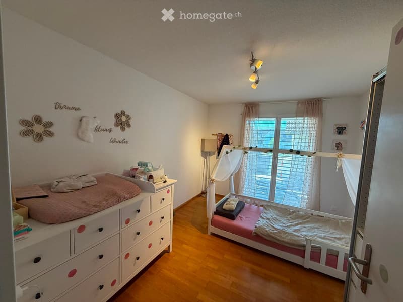 Cozy children's bedroom with a single bed, a changing table, and a window with curtains. The room features wooden flooring and decorative wall elements.