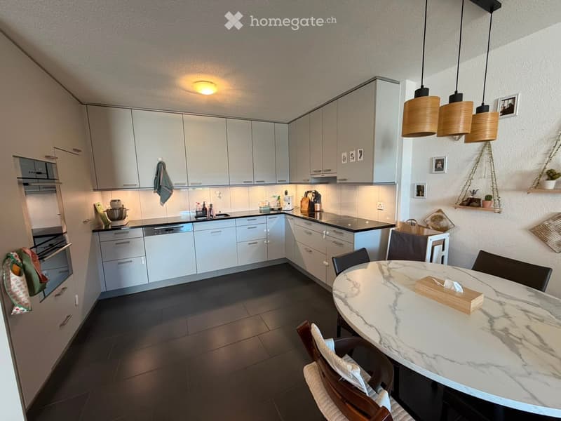 Modern kitchen with light grey cabinets, stainless steel appliances, a marble dining table with chairs, and pendant lights.