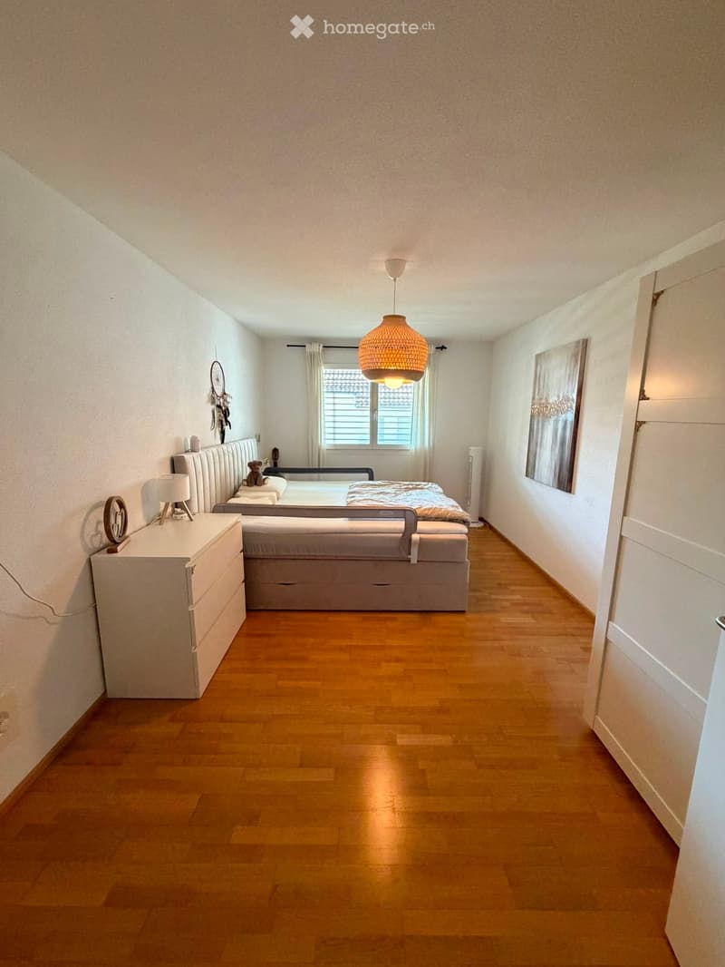 Modern bedroom with wooden flooring, a large bed with grey bedding, a nightstand with a lamp, a hanging woven lamp, and a window with curtains.