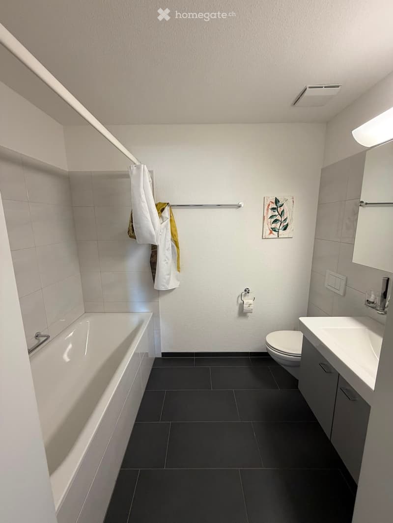 Modern bathroom with a bathtub, toilet, sink, and a towel hanging on a rack. The walls are tiled, and the floor is covered with dark tiles.