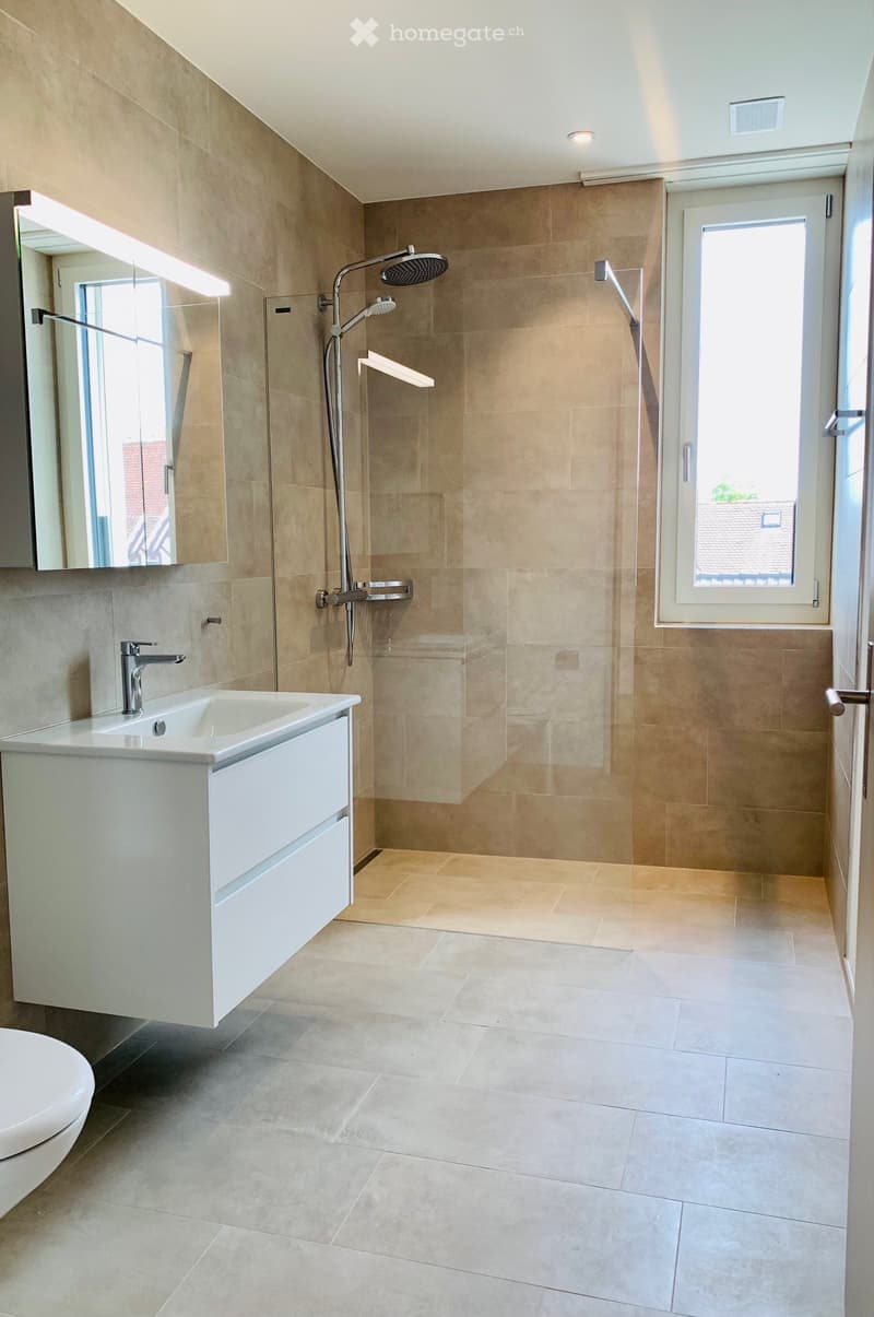 Modern bathroom with beige tiles, a white sink with a chrome faucet, a large mirror, a walk-in shower with a modern showerhead, and a toilet.