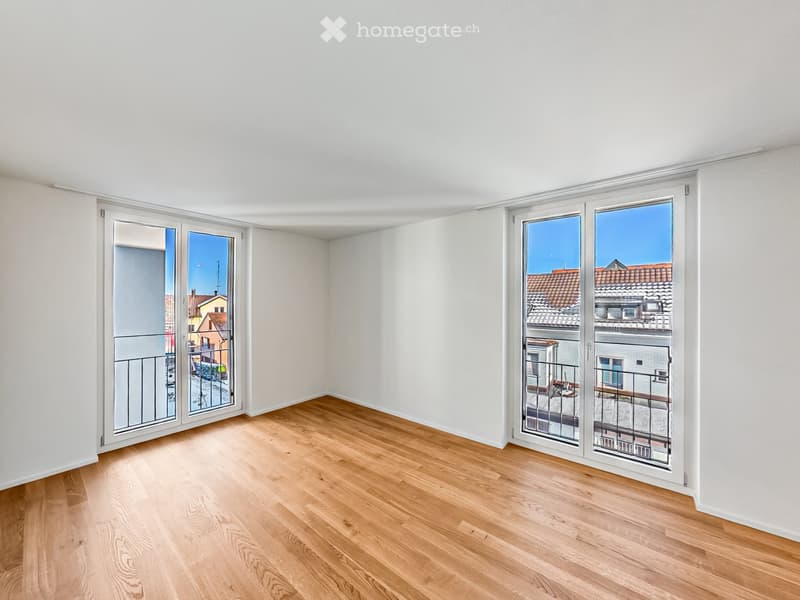 Empty room with light wooden flooring and large sliding glass doors leading to a balcony with a view of rooftops.