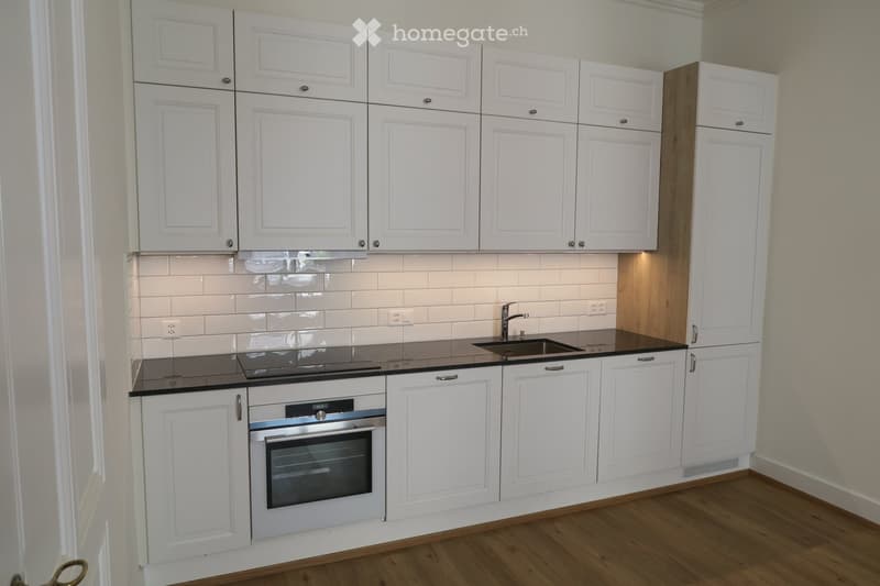 Modern kitchen with white cabinets, black countertops, and a built-in oven. Features a stainless steel sink and faucet.
