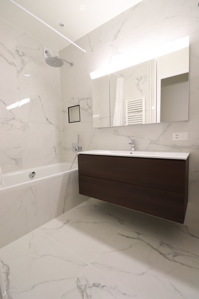 Modern bathroom with marble tiles, a bathtub, a sink with a dark wooden cabinet, a large mirror, and a showerhead.