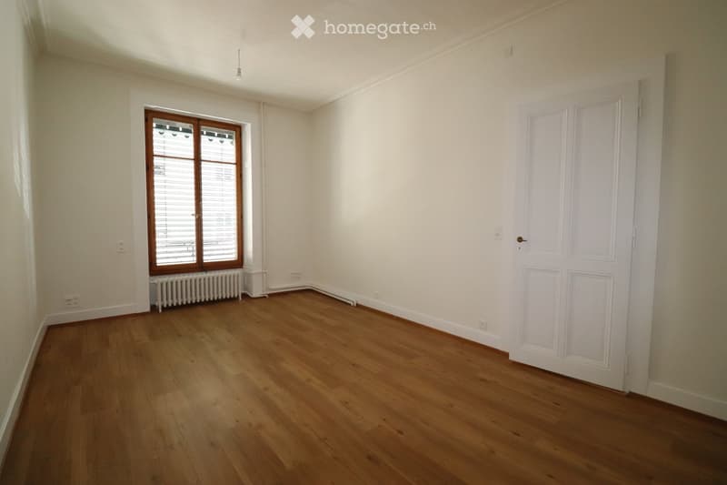 Empty room with wooden flooring, white walls, a window with blinds, a radiator, and a white door.