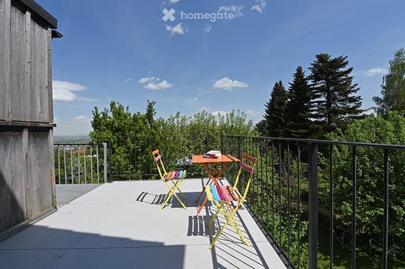Balcony with a small table and chairs, surrounded by a metal railing and lush greenery, under a clear blue sky.