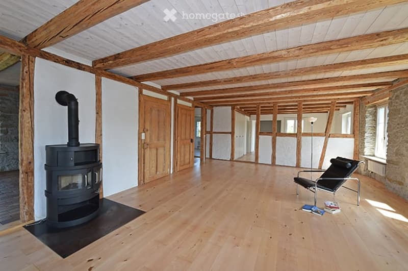 Open-plan space with exposed wooden beams, wooden flooring, and a black wood-burning stove. The room is unfurnished, with only a single black chair and a few books visible.