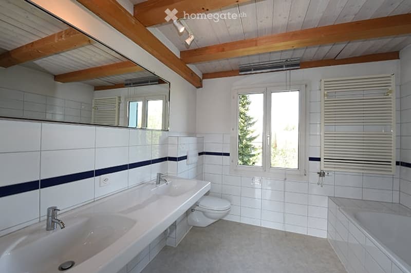 Bright bathroom with white tiled walls, two sinks, a toilet, a bathtub, and a large mirror. The room features wooden beams on the ceiling and a window with blinds.