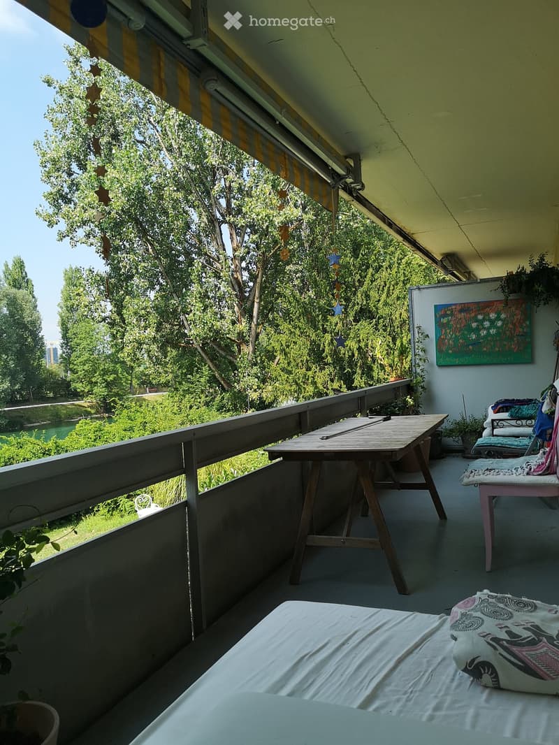 Balcony with a wooden table, chairs, a bed with white sheets, and a colorful painting on the wall. The balcony has a view of greenery and a river.