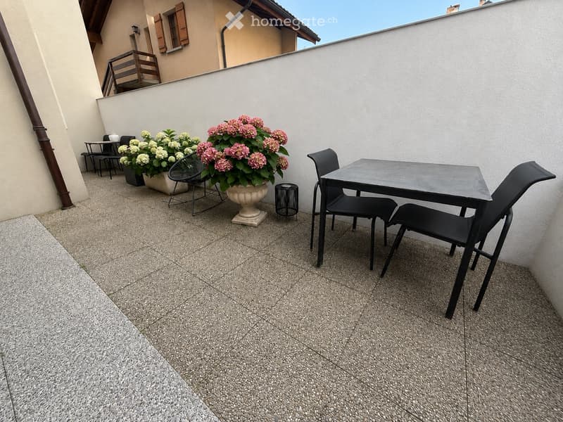 Balcony with a tiled floor, a small table with four black chairs, and two flower pots with pink and white flowers.