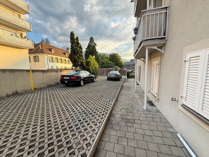 A narrow, paved alleyway between buildings with two parked cars, surrounded by trees and a distant view of mountains.