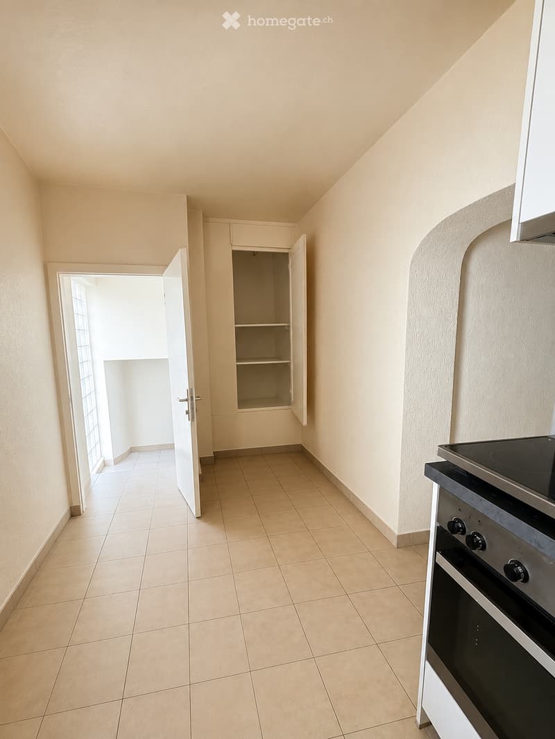 Empty kitchen with tiled floor, white walls, and a built-in oven on the right side. An open cabinet with empty shelves is visible in the background, and a doorway leads to another room.