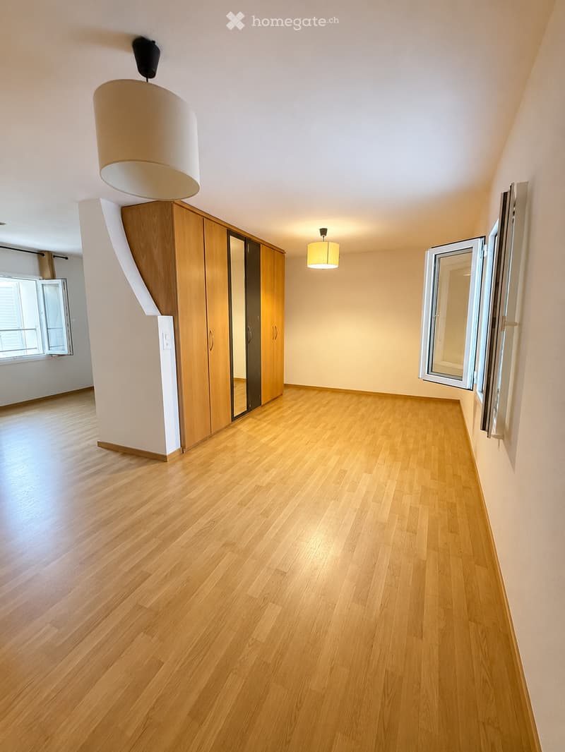 Empty room with wooden flooring, light-colored walls, and large windows. Features a built-in wooden wardrobe with mirrored doors and modern lighting fixtures.