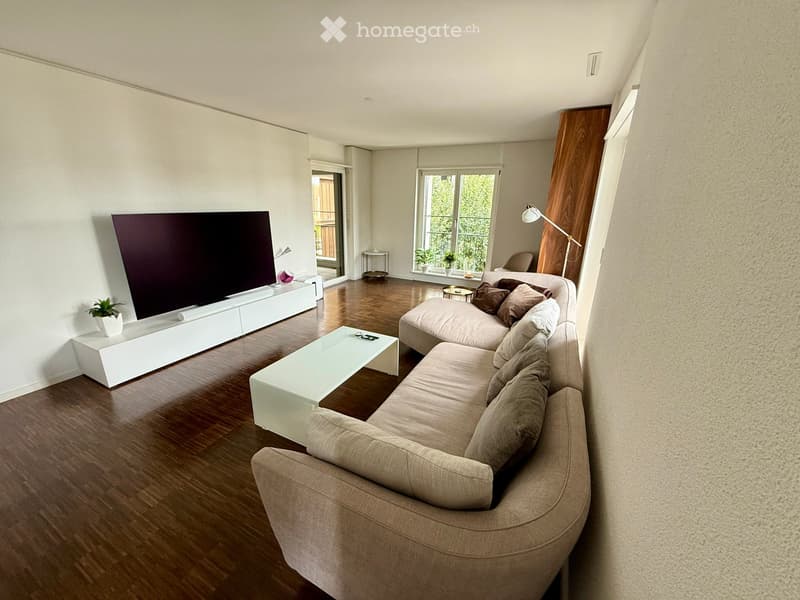 Modern living room with a large sectional sofa, coffee table, flat-screen TV on a white console, and a floor lamp near a large window with a view of a balcony.