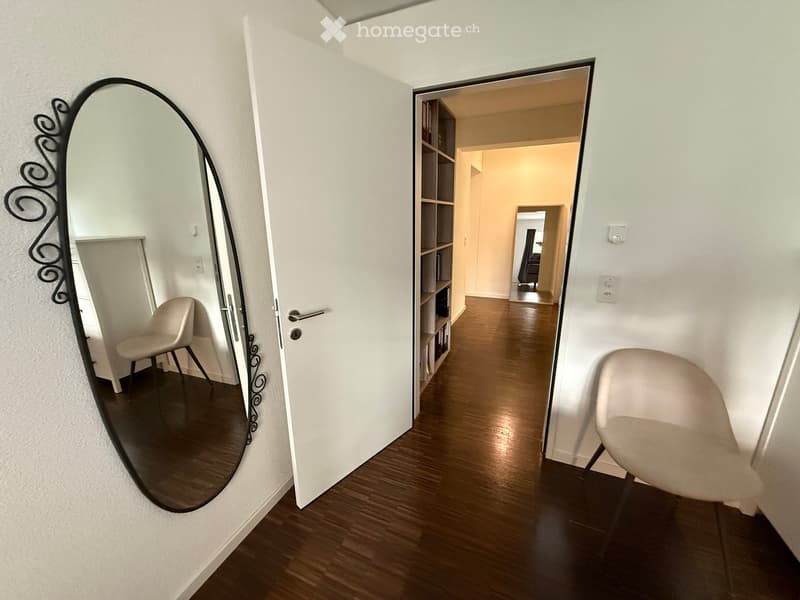A modern hallway with wooden flooring, white walls, and a large oval mirror on the left side. An open door leads to another room with a bookshelf and a chair visible in the background.