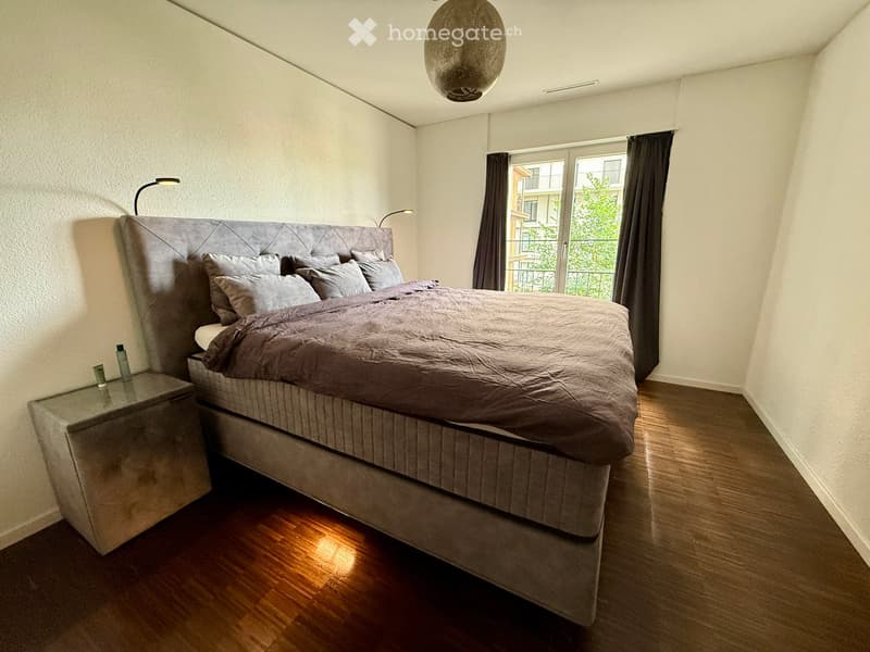 Modern bedroom with a large bed, nightstand, and a large window with curtains. The room features wooden flooring and a pendant light.