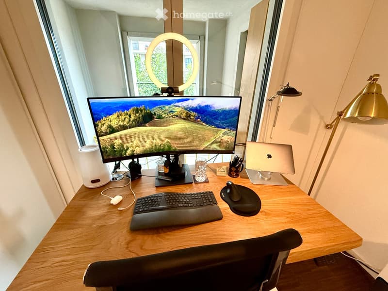 A modern, well-lit workspace with a wooden desk, ergonomic chair, large monitor, laptop, keyboard, mouse, and two lamps. The desk is organized with a cup, speaker, and other small items.
