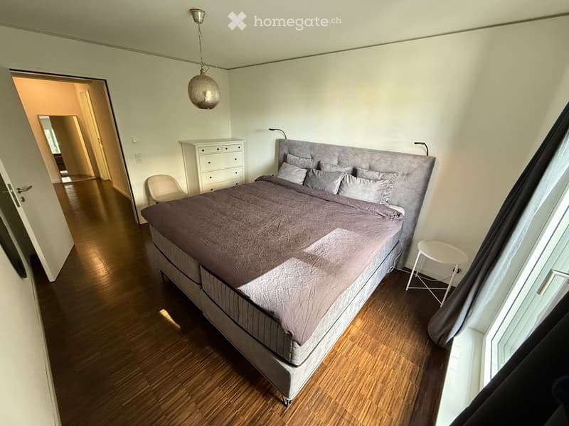 Modern bedroom with a large bed, nightstand, chair, and stool. The room features wooden flooring and large windows with curtains.