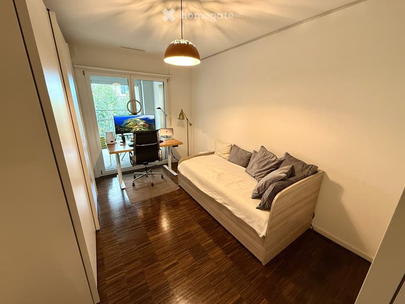 A small, modern furnished room with a daybed, desk, and chair. The room features wooden flooring, a hanging pendant light, and a large window with a view of greenery.