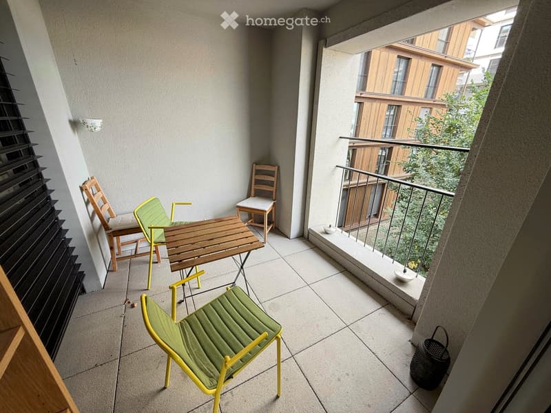 Balcony with tiled floor, wooden table, green chairs, and a small stool, overlooking a green area with trees.