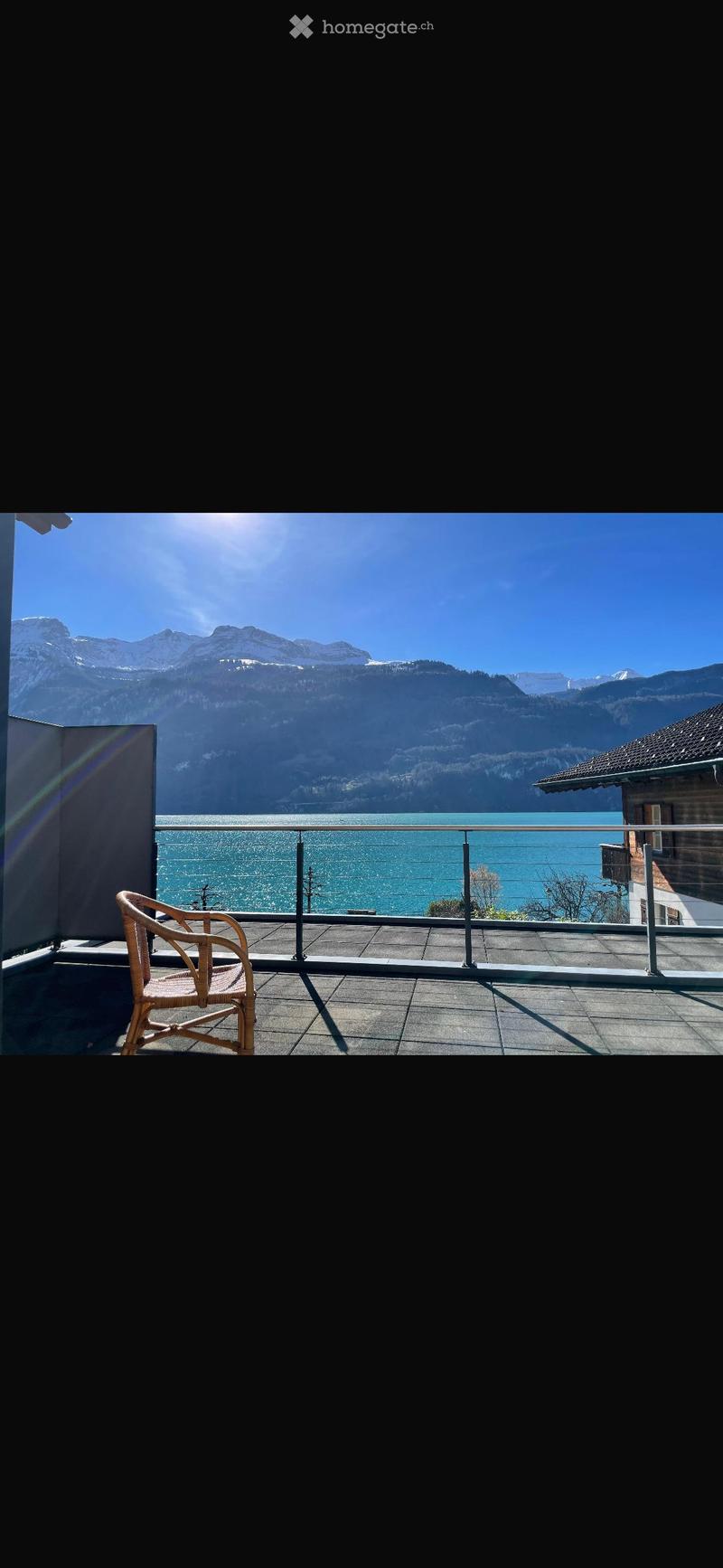 Balcony with a view of a lake and mountains, featuring a single wicker chair and a metal railing.