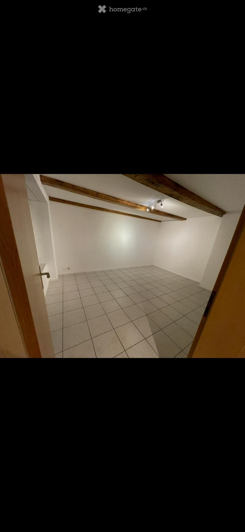 Empty room with tiled floor, white walls, wooden beams on the ceiling, and a single light fixture.