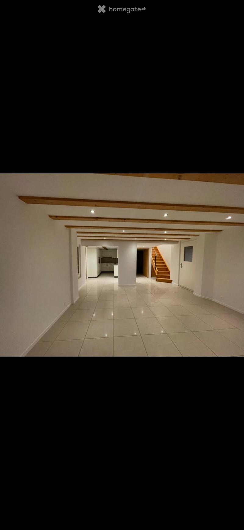 Empty hallway with tiled floor, white walls, wooden beams on the ceiling, and visible rooms including a kitchen and staircase in the background.