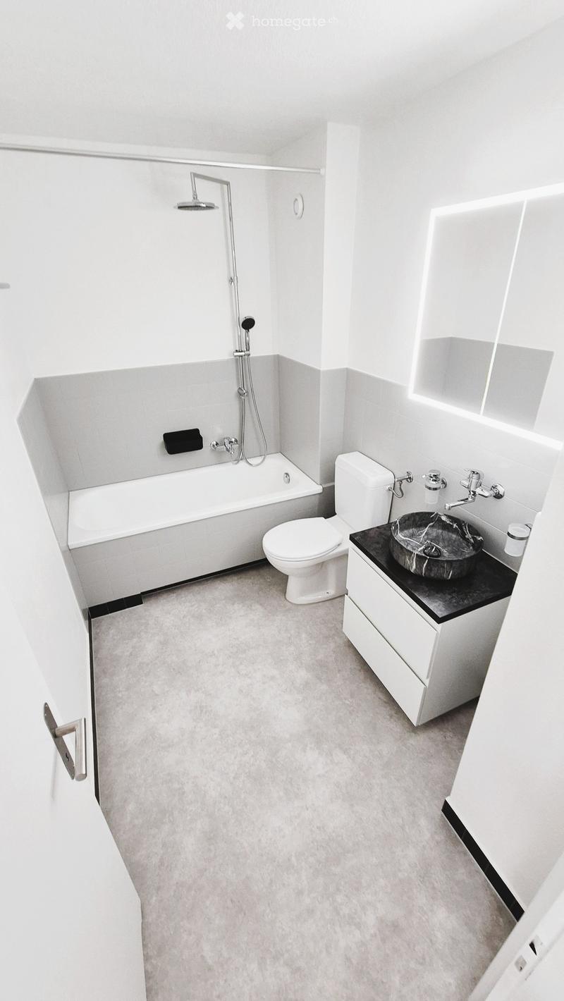 Modern bathroom with a bathtub, toilet, sink, and shower head. Features a sleek, minimalist design with white tiles and a grey countertop.