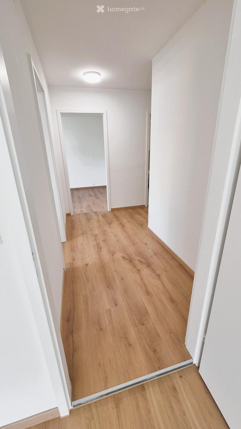 Empty hallway with light wooden flooring and white walls, featuring a recessed ceiling light and an open doorway leading to another room.
