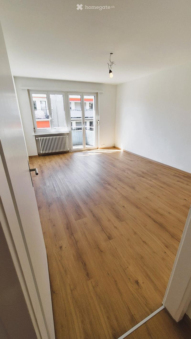 Empty room with wooden flooring, white walls, large window with sliding glass door, radiator, and a single hanging light fixture.