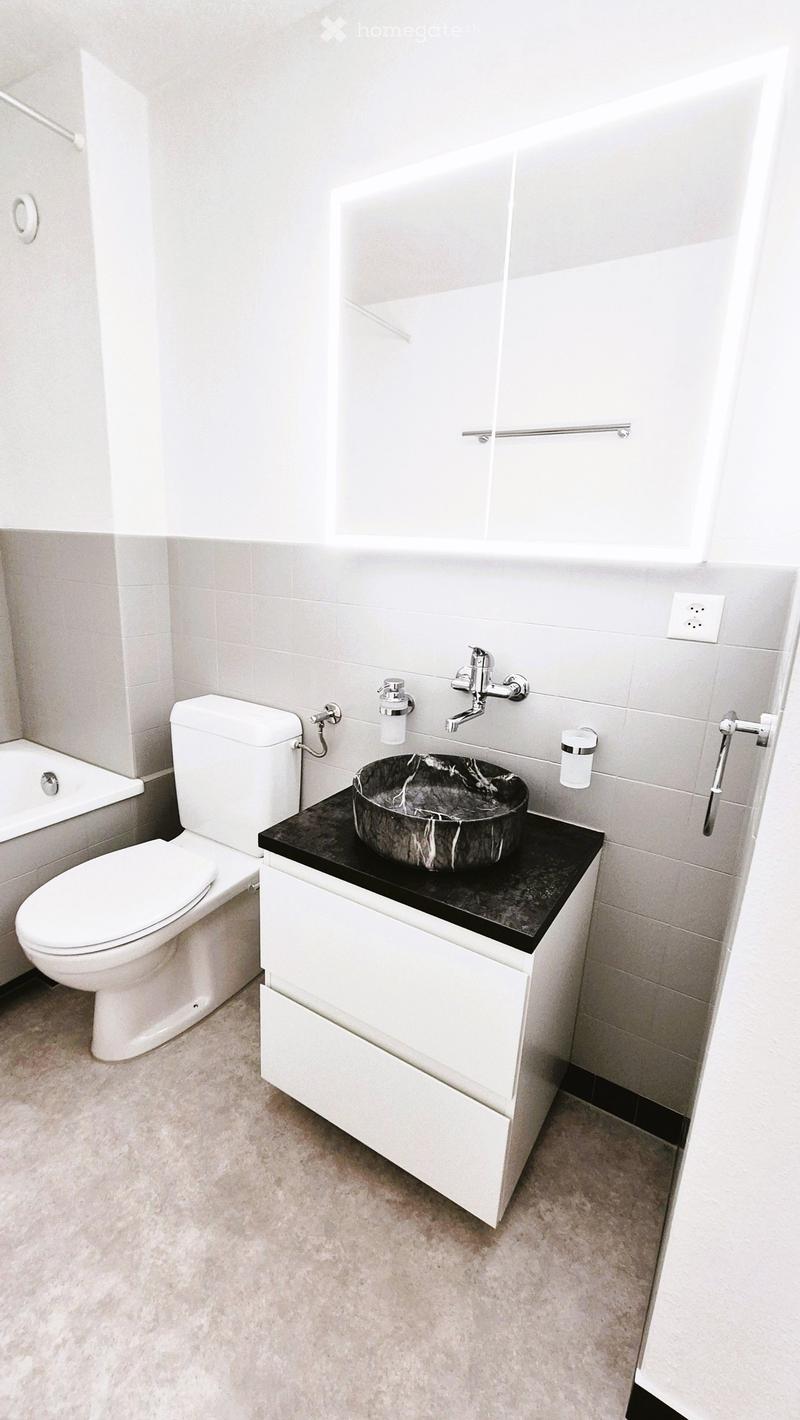 Modern bathroom with a white toilet, black countertop sink, chrome faucet, and a bathtub. The walls are tiled in light grey, and the floor is covered with beige tiles.