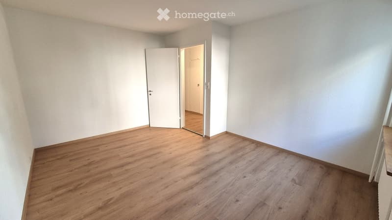Empty room with light wooden flooring and white walls, featuring an open door leading to another room.
