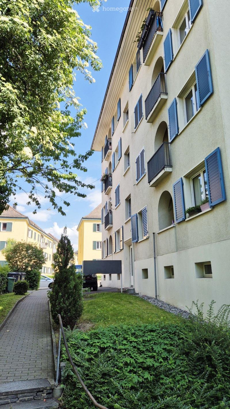Multi-story residential building with light yellow facade, blue shutters, and multiple balconies. The building is surrounded by greenery and a paved pathway.