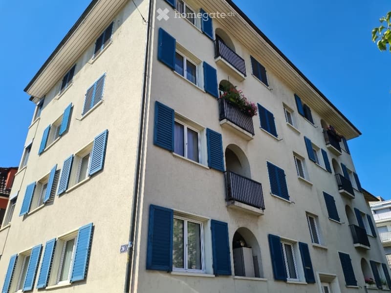 Four-story building with light beige facade, blue shutters, and multiple balconies with flower boxes.