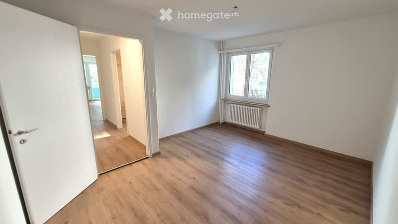 Empty room with wooden flooring, white walls, and a window with a radiator below it. The room has an open doorway leading to another area.