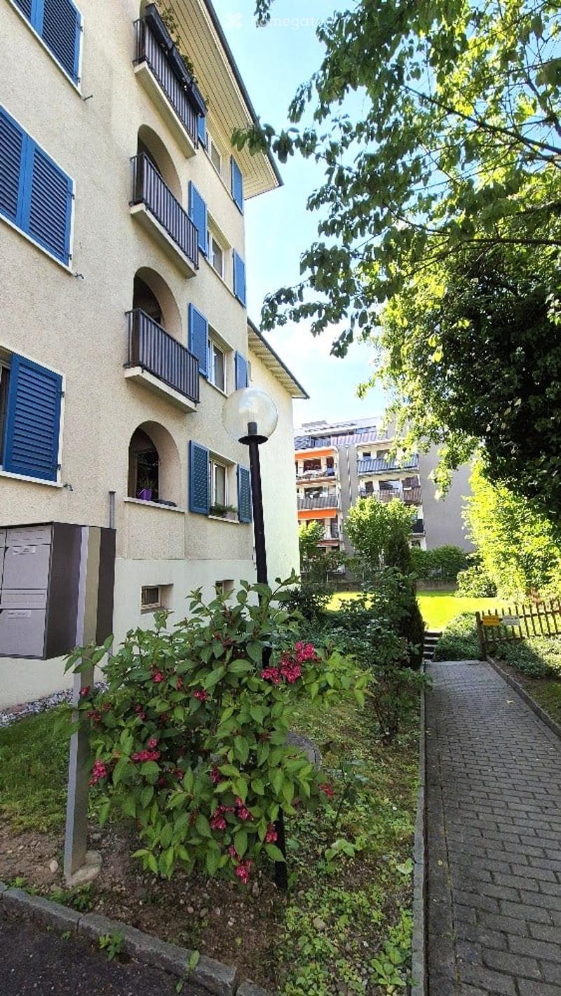 Multi-story residential building with blue shutters, balconies, and a small garden area with flowers and a lamp post.