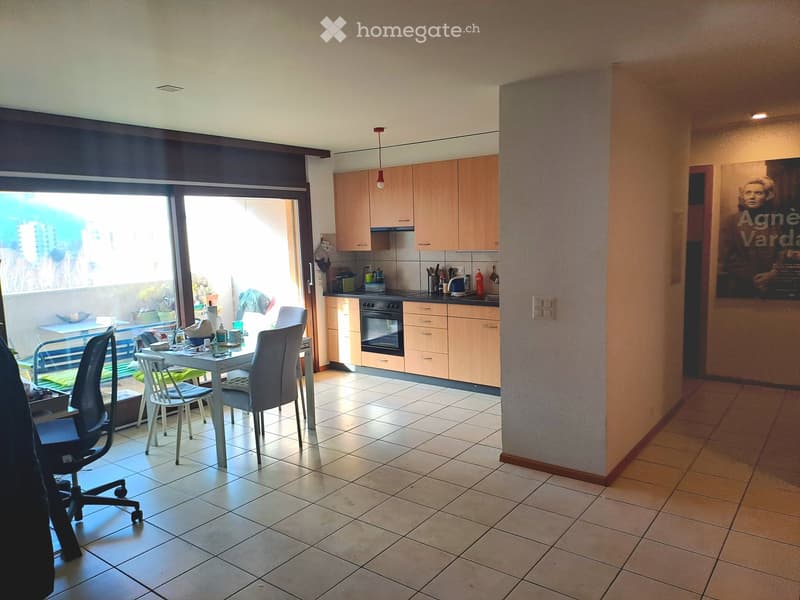 A modern kitchen and dining area with light wooden cabinets, a white dining table with four chairs, and a view to a balcony. The kitchen features an oven, refrigerator, and various small appliances.