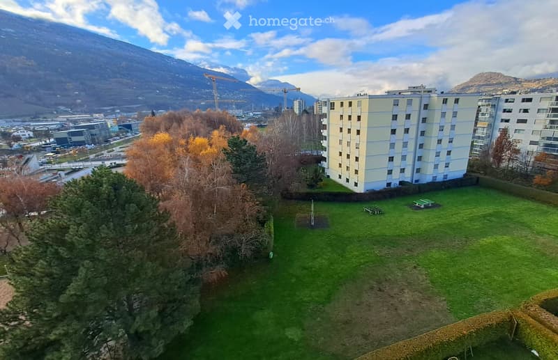 A grassy outdoor area with trees, a bench, and a view of a multi-story building and distant mountains.