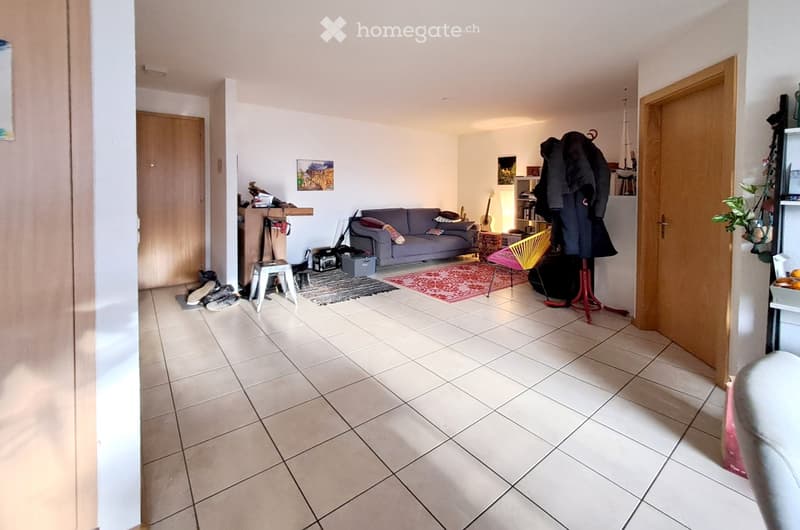 Living room with tiled floor, grey sofa, small stool, guitar, coat rack, plant, and a small table with a chair.