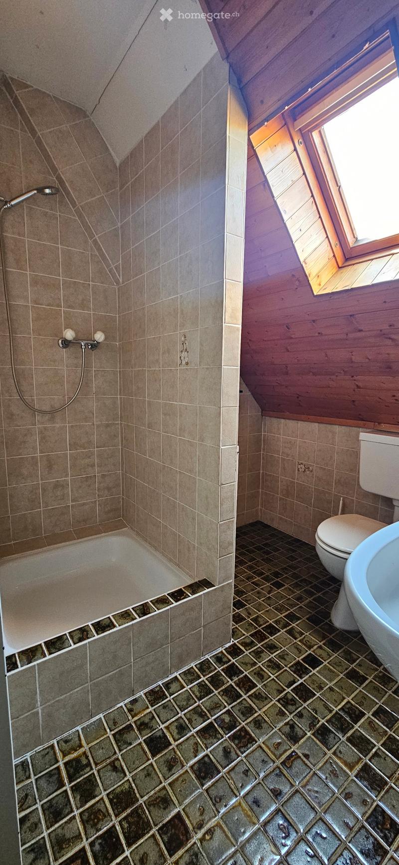 Bathroom with a bathtub, toilet, sink, and a window with wooden frame. The walls are tiled in beige and the floor is tiled in dark brown.