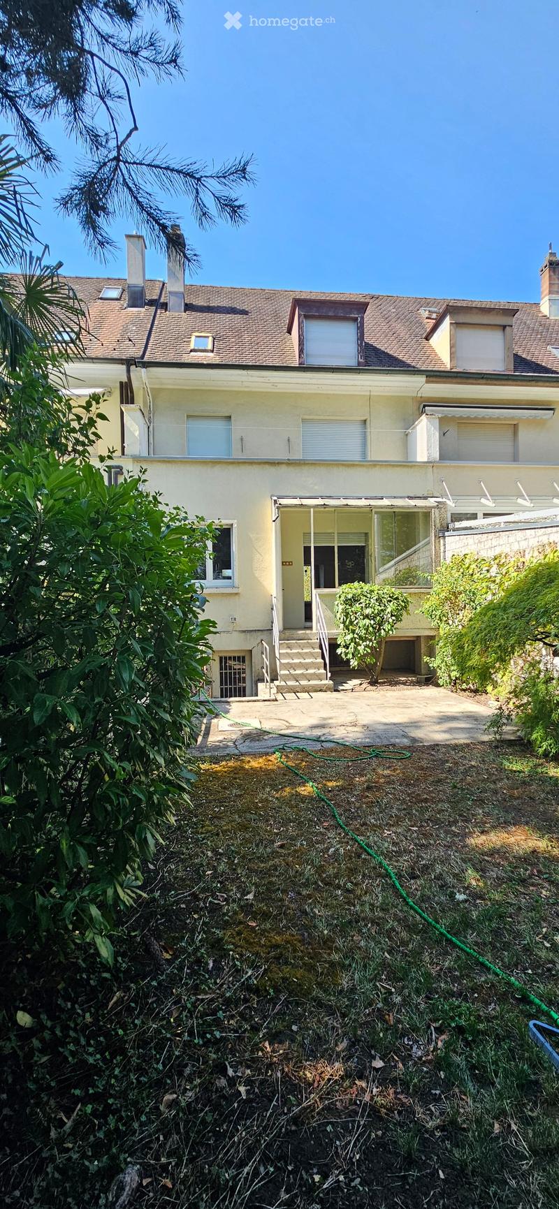 Two-story house with beige exterior, multiple windows, and a covered entrance leading to a small patio area. The yard is partially overgrown with grass and plants.