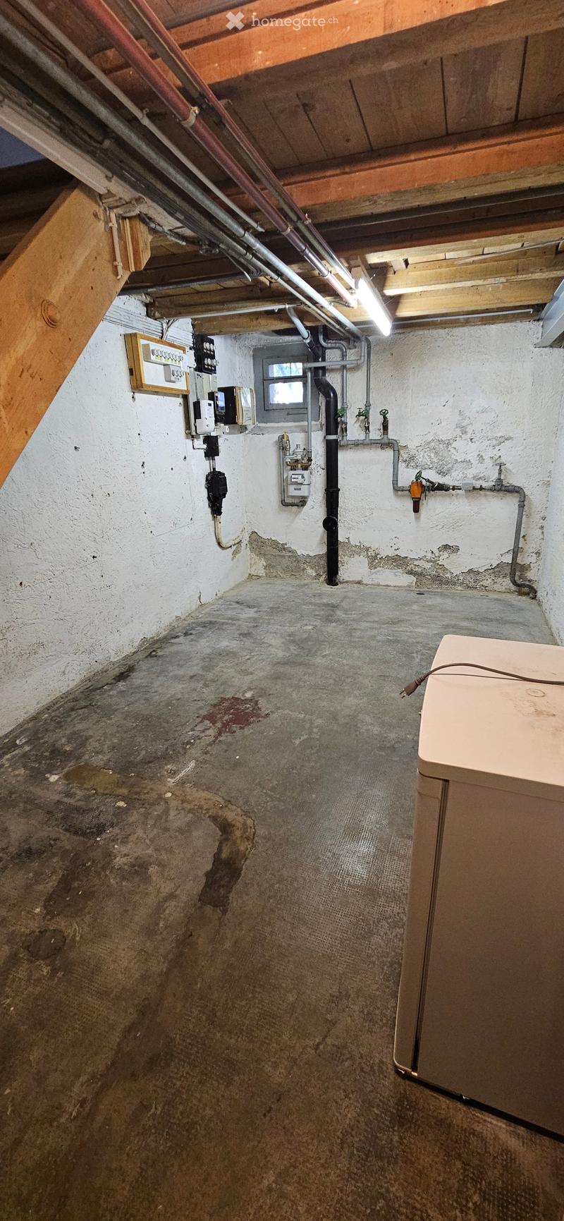 Empty basement space with exposed wooden beams, electrical panels, and plumbing pipes. Concrete floor with visible stains and wear.