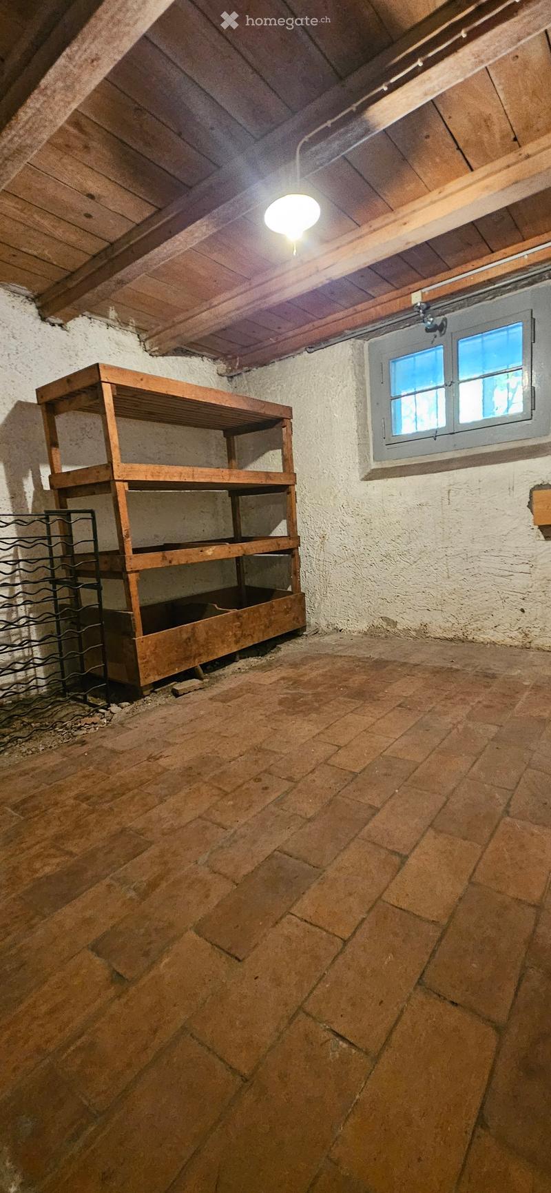 Empty room with exposed wooden beams, stone walls, and a wooden shelf. The floor is made of large stone tiles, and there is a small window with frosted glass panes.