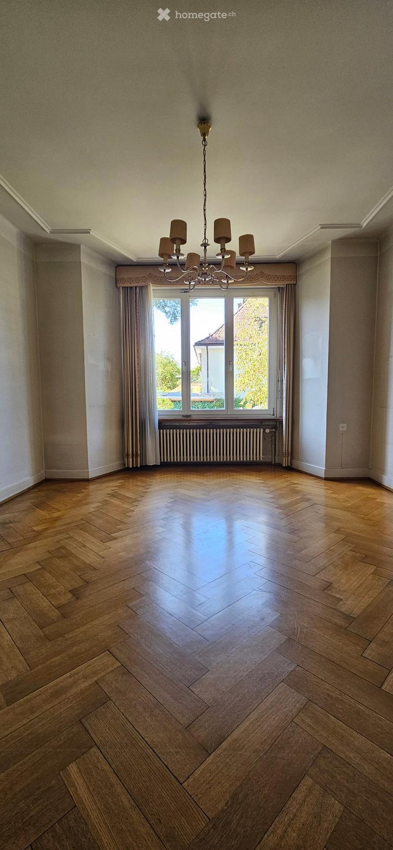 Empty room with wooden herringbone flooring, large window with curtains, radiator, and a chandelier.