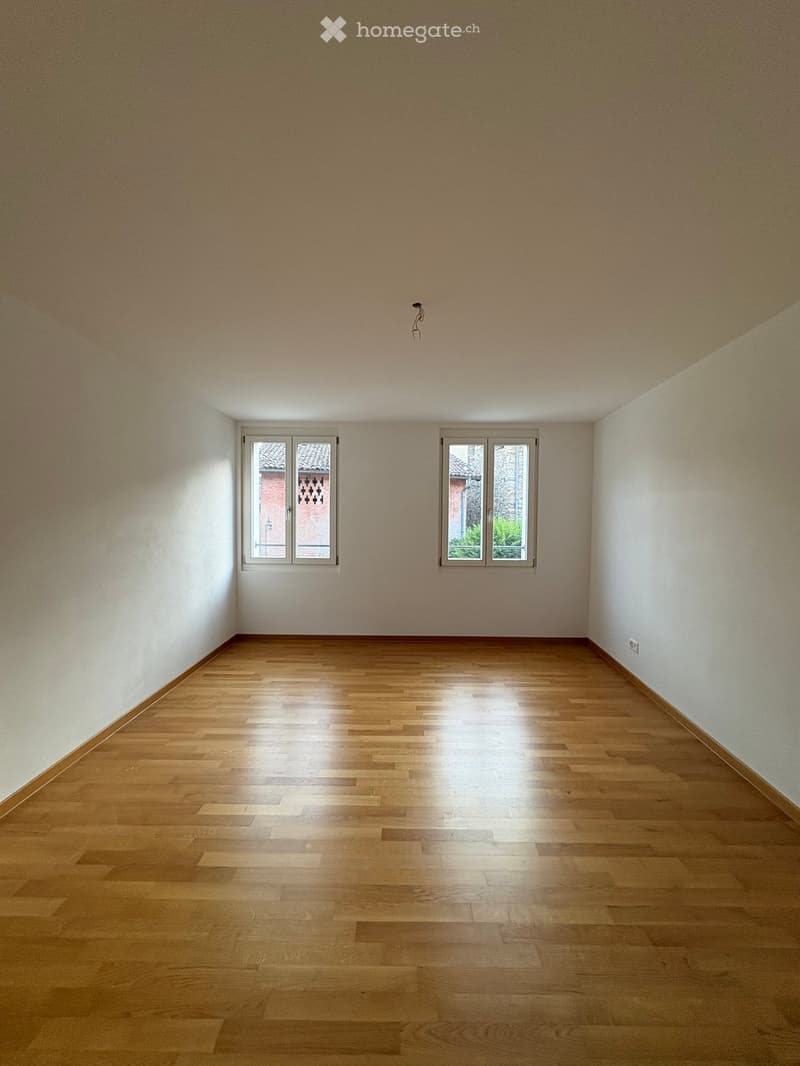 Empty room with wooden flooring and white walls, featuring two windows with white frames and a visible electrical outlet on the right wall.