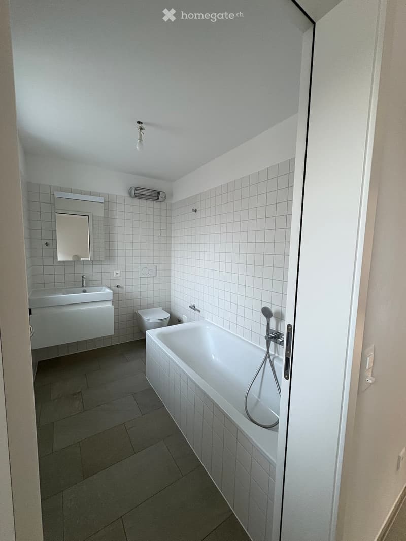 Modern bathroom with white tiled walls, a bathtub, toilet, sink, and mirror. The space features a sleek design with a built-in showerhead and a modern light fixture.