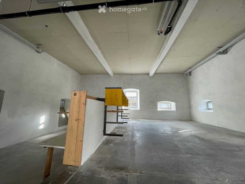 Empty room with concrete floors and walls, exposed pipes, and minimal lighting. Features a small window and a few built-in shelves.