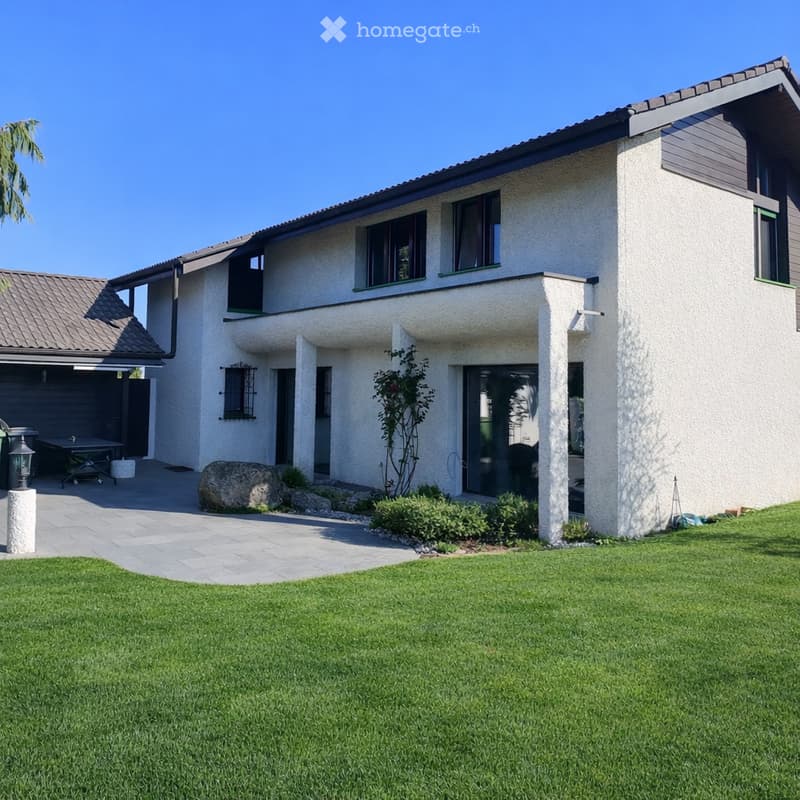 Two-story house with white stucco exterior, dark green trim, and a modern design. Features include a garage, a patio area with a table and chairs, and a well-maintained lawn.